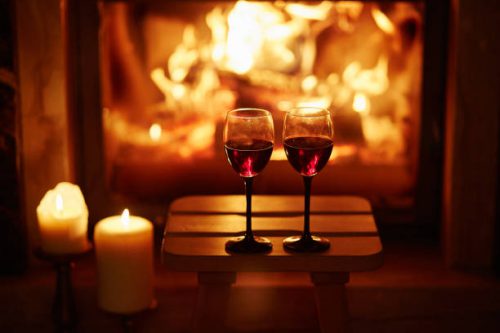 Two glasses of red wine near fireplace with many candles. Cozy romantic evening for couple or Christmas celebration concept